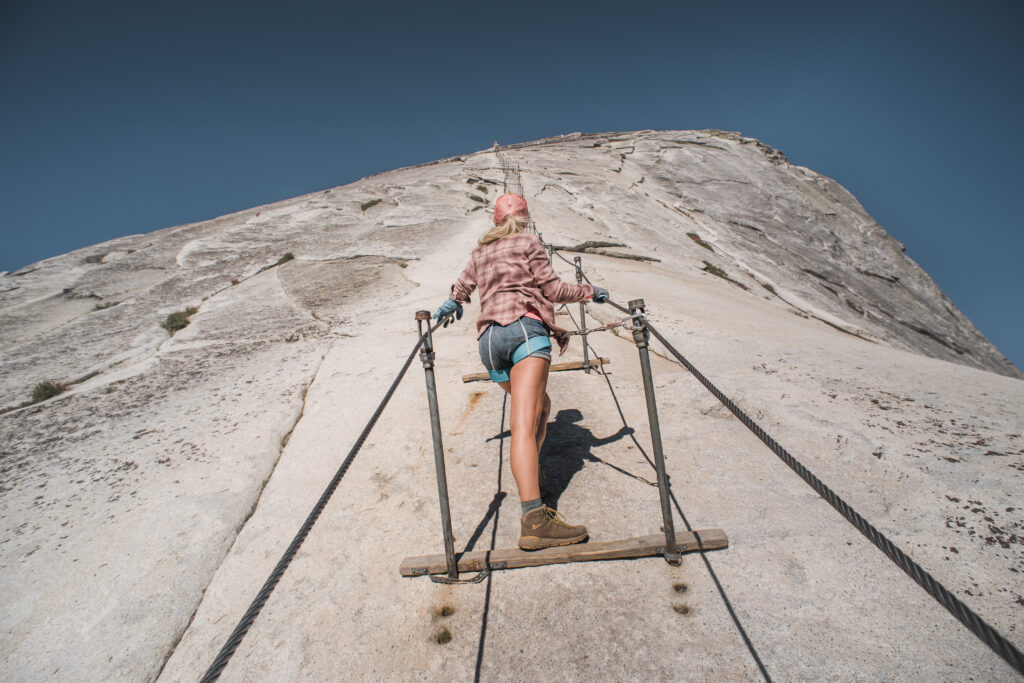 A woman stands looking up on the half dome cables wearing a harness