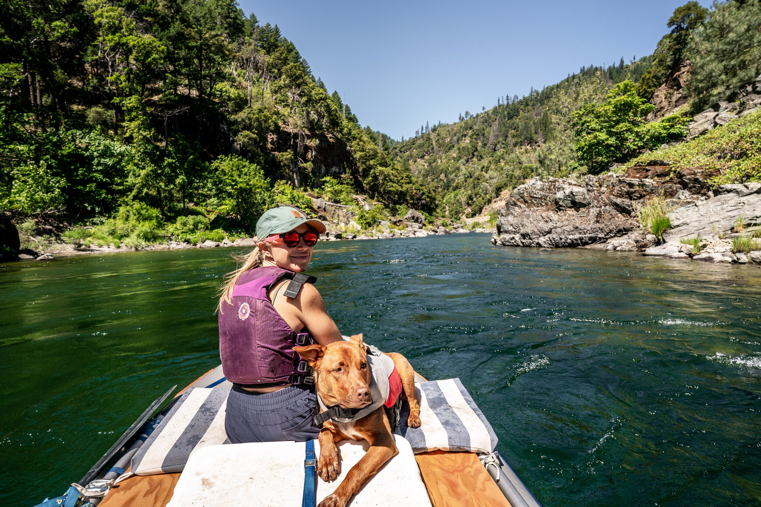 A woman and a dog sit on a raft, wearing lifejackets on a river