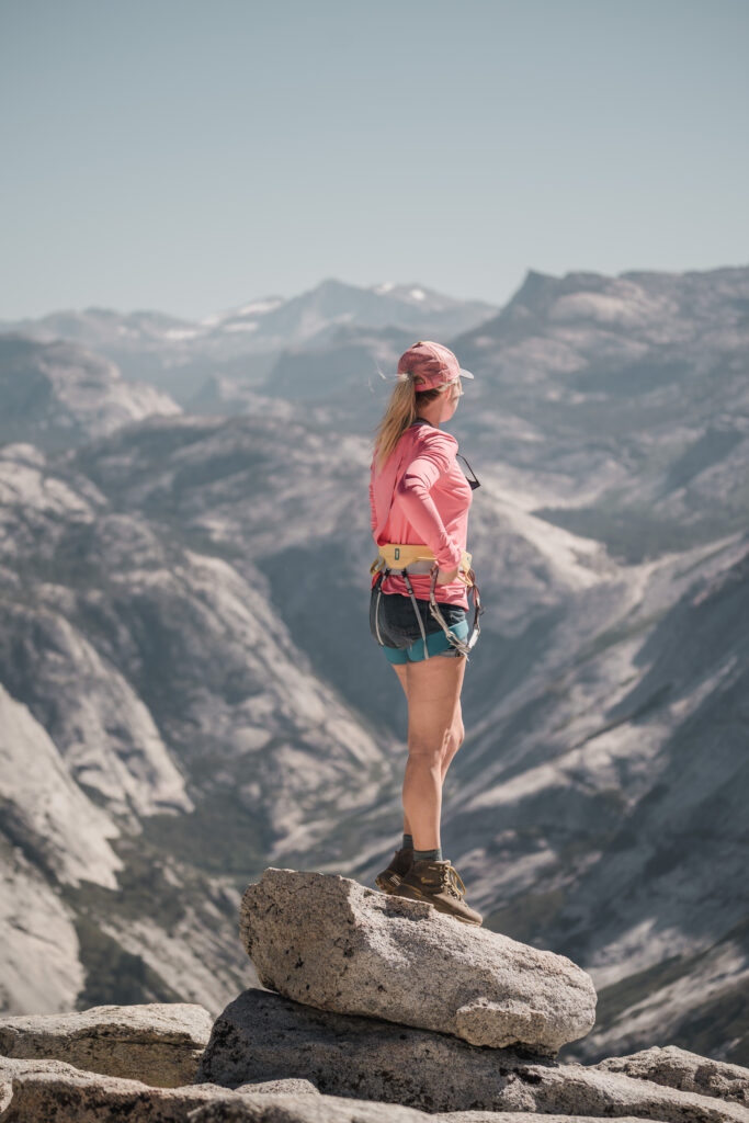A woman stands on top of Half Dome looking out while wearing a climbing harness and hiking gear at Yosemite National Park