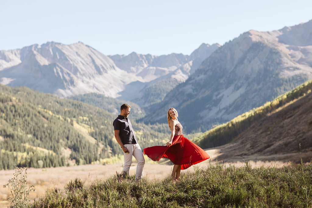 A woman twirls wearing a red dress for her fiancé who looks on at Pine Creek Cookhouse in Aspen Colorado. They are standing in front of the Elk Mountain Range