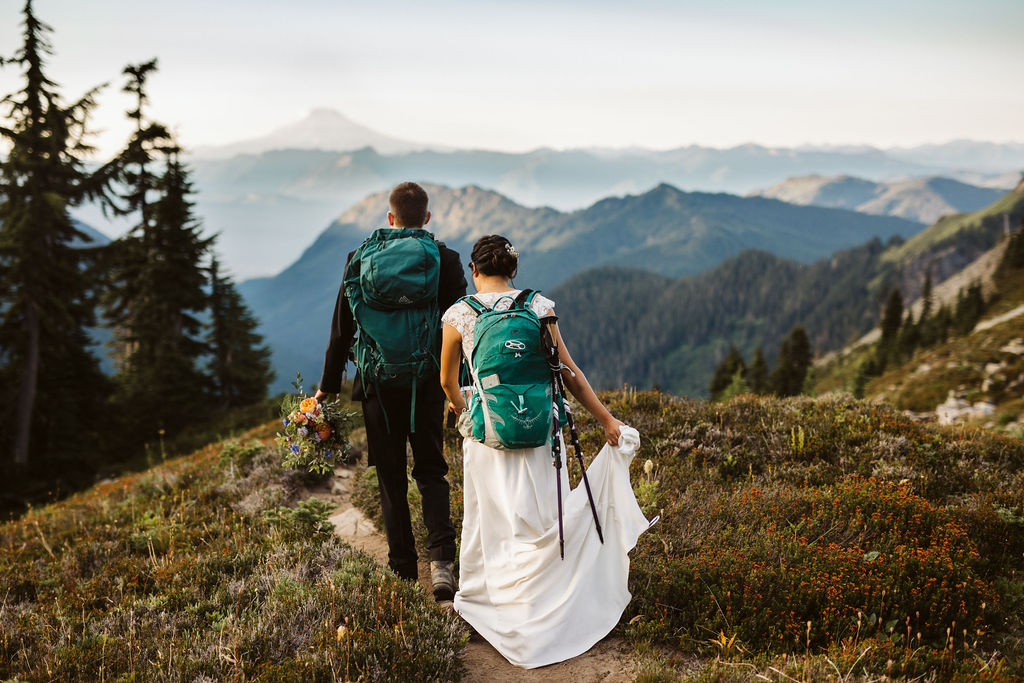 a bride and groom wearing backpacks hike along a trial in the pacific north west for an adventure elopement photoshoot at sunset